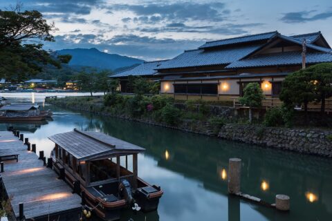 Pleasure boat and Togetsu Café old building with yellow lights at sunset, on the river bank of Saganakanoshimacho island and Arashiyama Park Nakanoshima area, Katsura River and Arashiyama hills, Ukyo Ward, Kyoto, Japan.