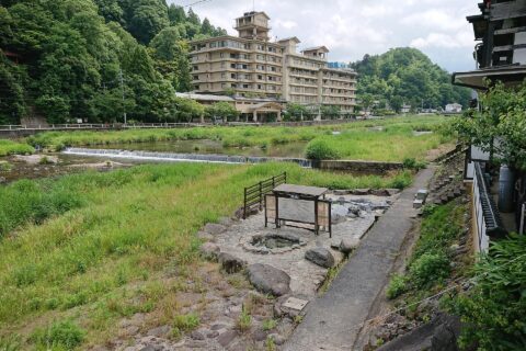 鳥取県三朝町の三朝温泉、河原風呂と三徳川。三朝橋のたもとから上流方向を撮影しており、手前が足湯「河原の湯」、屋根のある部分が脱衣所でその奥が「河原風呂」の湯船。入湯は無料。右側が温泉街として旅館が密集しているエリアで、写真正面は旅館「依山楼 岩崎」、中央右奥にキュリー広場（川に背を向ける形で屋根付き舞台がある）、右奥に恋谷橋が見える。